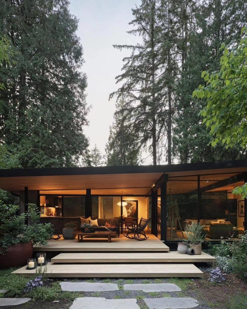 Mid-century modern house with glass walls and wooden deck surrounded by tall trees.