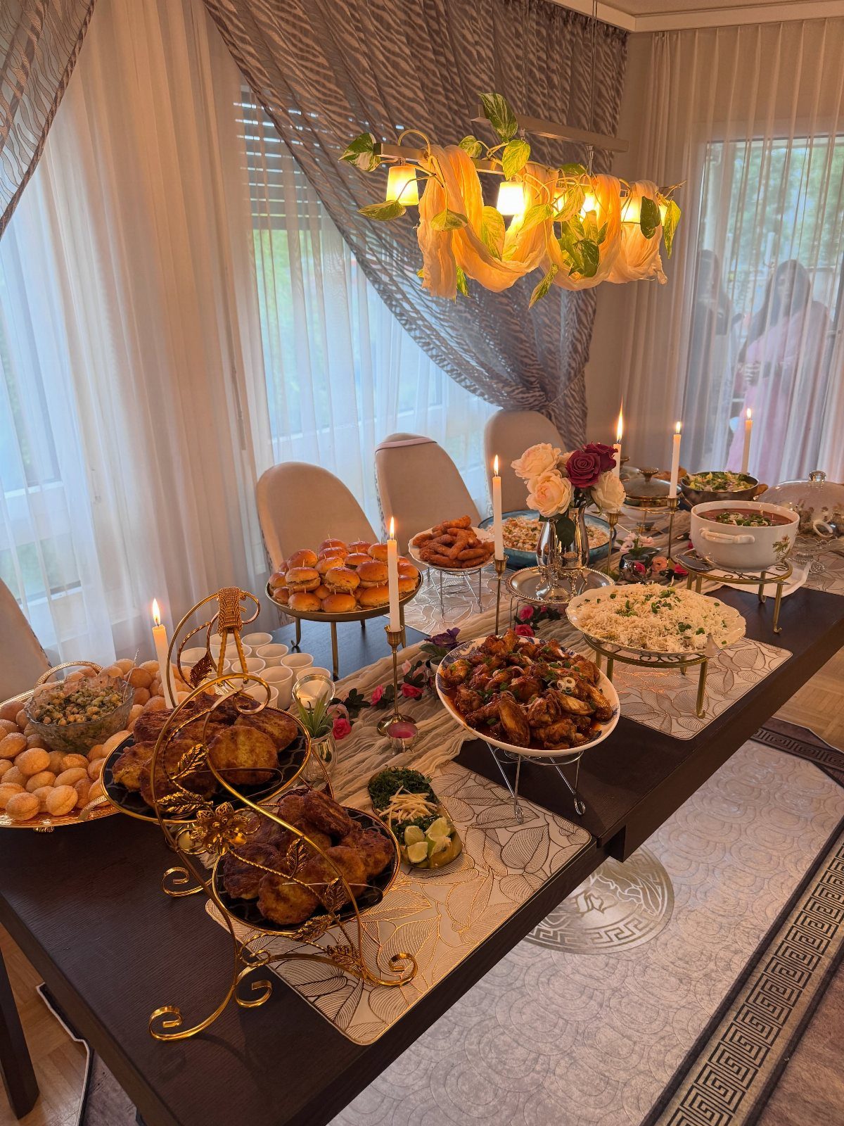 Dinner table decorated with food platters, white candles, and flowers under a yellow chandelier.
