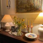 Wooden sideboard with lamps, yellow wildflowers, and a gold-framed landscape painting above.