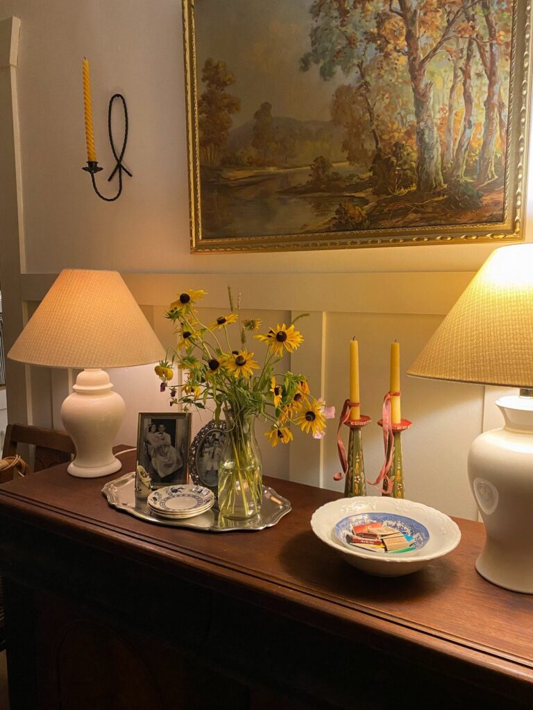 Wooden sideboard with lamps, yellow wildflowers, and a gold-framed landscape painting above.