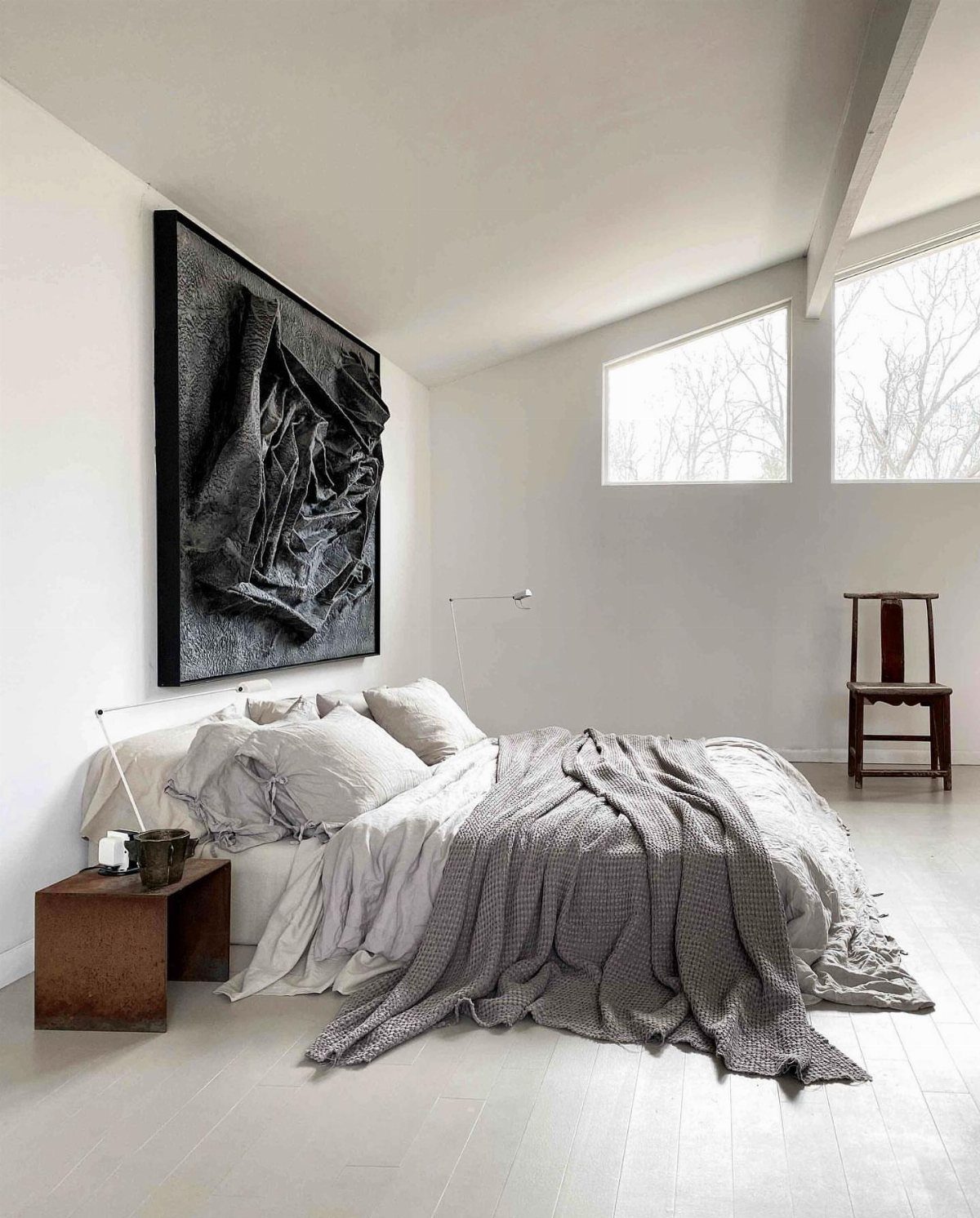 Minimalist white bedroom with grey bedding and a large black textured wall art.